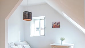 Une pièce avec un plafond en pente, un lit avec une literie blanche, une table blanche avec deux tabourets, un luminaire suspendu et une fenêtre.