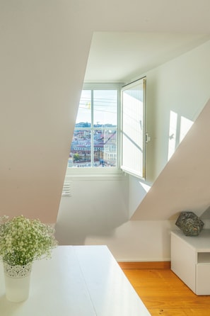 A room with a sloped ceiling, a window with a view of a cityscape, a white table, and a small white cabinet.