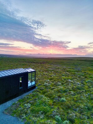 A modern building with a glass facade, situated in a grassy field with a distant mountain range and a colorful sky. - Tiny Glass lodge Experience The Nature (Selfoss)