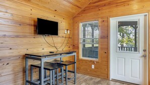 A wooden-paneled room with a television mounted on the wall, a window with blinds, and a white door. - Pine Lakes Adventure Base | Swim, Fish & Explore the Great Outdoors (Pike County)
