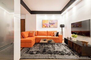 A modern living room with a sectional sofa, a coffee table, a black rug, a flat-screen TV, and a painting on the wall. - Acropolis Exclusive Apartment | 34 Dimitrakopoulou str 11742 Athens (Athens)