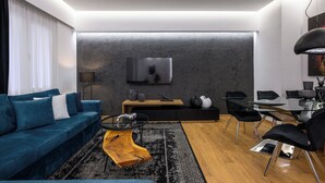 Un salon moderne avec un canapé bleu, une table basse en verre, une table d’appoint en bois, une table à manger noire avec des chaises, et un téléviseur à écran plat fixé au mur.