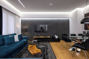 Ein modernes Wohnzimmer mit einem blauen Sofa, einem Glastisch, einem Holznachttisch, einem schwarzen Esstisch mit Stühlen und einem an der Wand befestigten Flachbildfernseher.