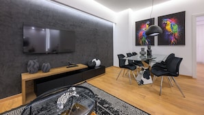 Un salon moderne doté d’un téléviseur à écran plat, d’un coin repas avec une table en verre et des chaises noires, ainsi que de deux œuvres d’art murales aux couleurs vives.