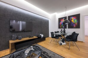 Ein modernes Wohnzimmer mit einem Flachbildfernseher, einem Essbereich mit einem Glastisch und schwarzen Stühlen sowie zwei farbenfrohen Wandbildern.