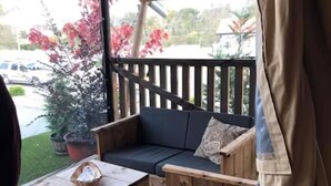 Un área de estar al aire libre con techo, mobiliario de madera, una mesa y vistas a la calle y árboles en floración de color rosa.