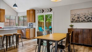 A modern kitchen with wooden cabinets, a dining area with a wooden table and chairs, and a view of the outdoors through a glass door.