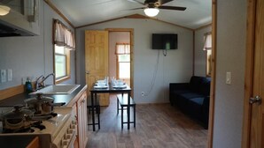Une cuisine compacte équipée d’une cuisinière, d’un évier et de meubles en bois, une table à manger avec des chaises, un ventilateur de plafond et un téléviseur fixé au mur.