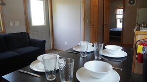Une table à manger dressée avec des assiettes, des bols et des verres, un canapé noir et un coin cuisine équipé d’un extincteur.