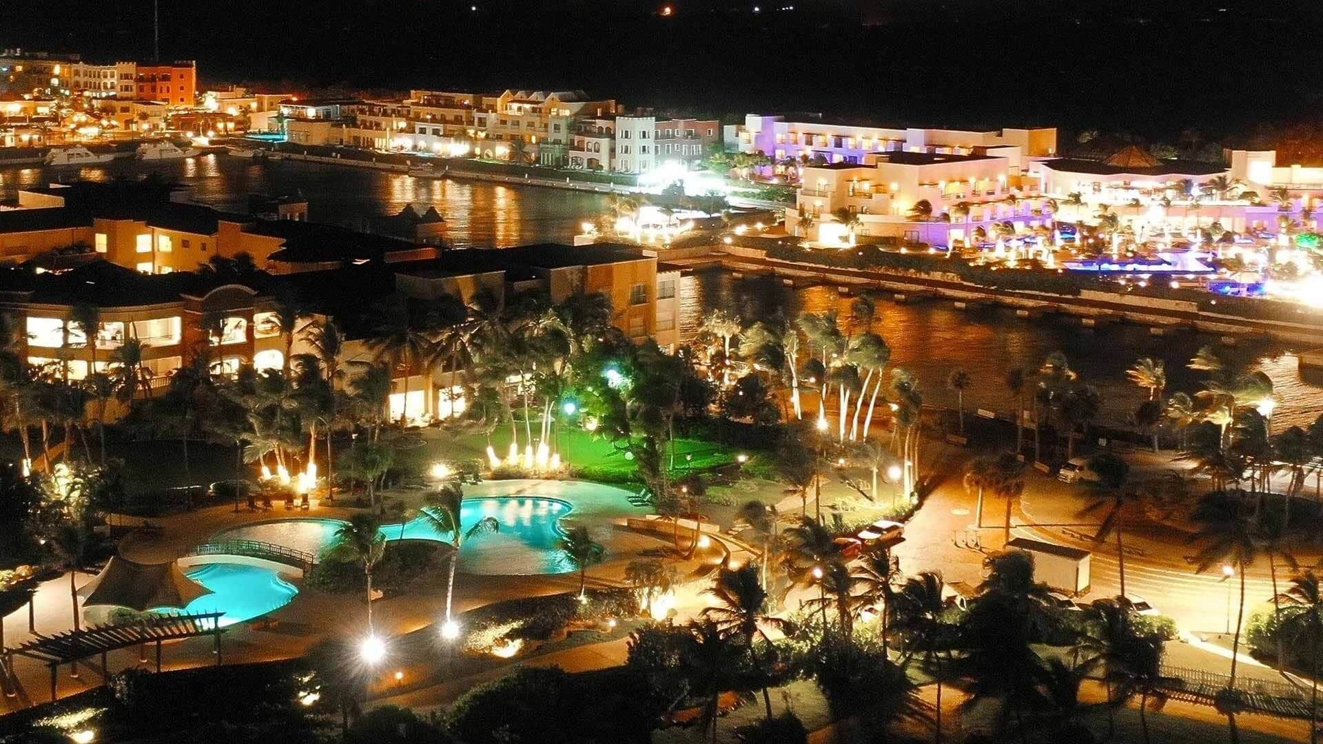 A resort with multiple swimming pools, palm trees, and illuminated buildings at night.