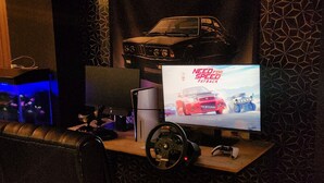 A gaming setup with a monitor displaying a racing game, a racing wheel, and a car model on the desk.