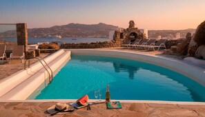 A swimming pool with a stone border, lounge chairs, and a bottle of wine on the edge. - Kanalia Home With Panoramic Views (Mykonos)
