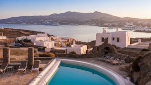 A pool area with lounge chairs, a stone wall, and white buildings with a view of the sea and mountains. - Kanalia Home With Panoramic Views (Mykonos)