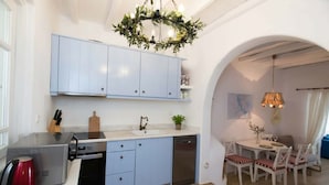 A compact kitchen with light blue cabinets, a dishwasher, a coffee maker, and a dining area with a table and chairs. - Kanalia Home With Panoramic Views (Mykonos)