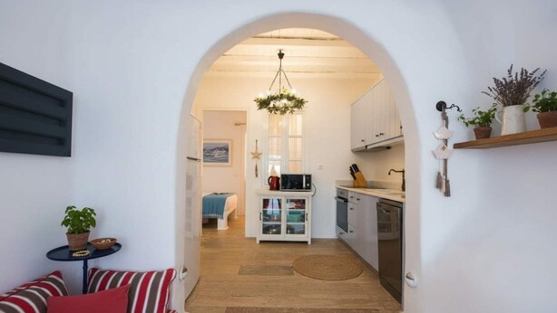 A narrow hallway leading to a kitchen with white cabinets, a microwave, and a hanging light fixture. - Kanalia Home With Panoramic Views (Mykonos)