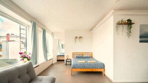Een slaapkamer met een bed, een bank, een kleine tafel, een raam met gordijnen en een wandplank met planten.