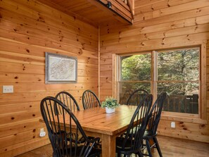 A wooden interior with a dining table, chairs, a framed map, and a window with a view of trees.