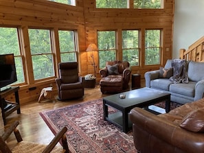 A wooden cabin interior with a large window, a television, leather furniture, and a patterned rug.