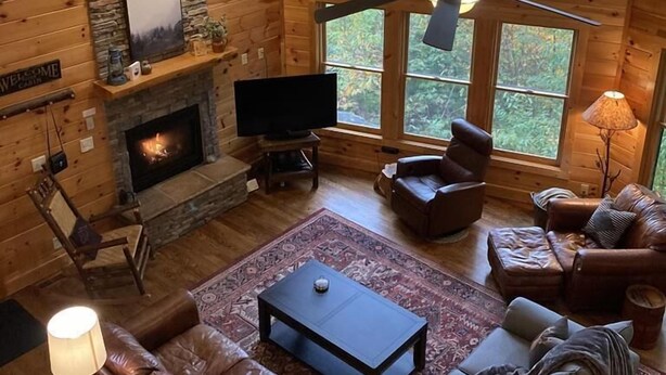 Ein gemütliches Blockhaus-Interieur mit Kamin aus Stein, Ledersofas, einem Fernseher und großen Fenstern mit Blick auf den Wald.