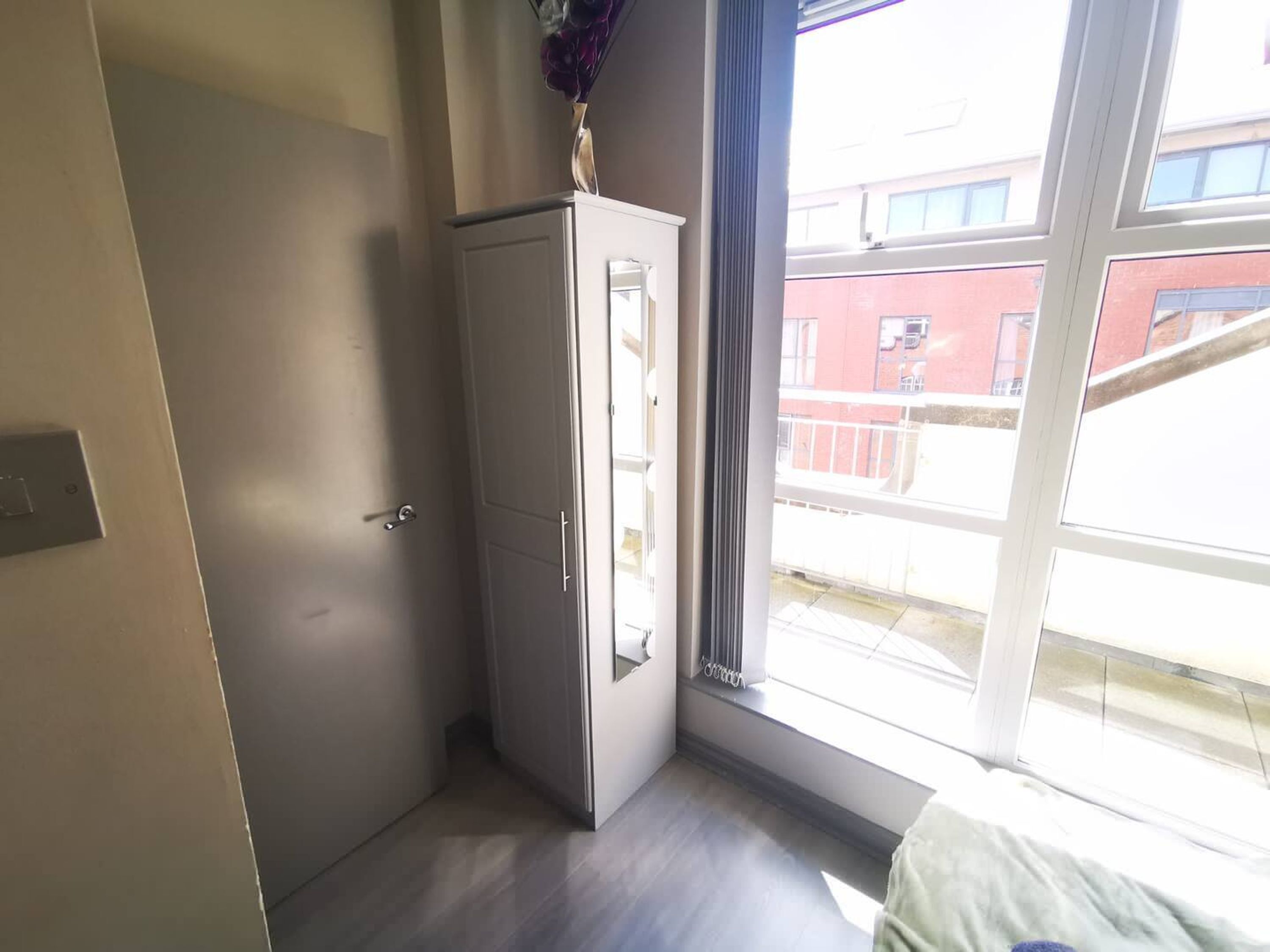 Ett rum med en vit garderob, en säng, ett fönster med utsikt över byggnader och en dörr.