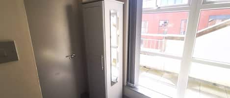 Ein Zimmer mit weißem Kleiderschrank, einem Bett, einem Fenster mit Blick auf Gebäude und einer Tür.