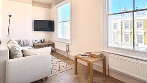 A living room with a sofa, a TV, a table with magazines, and a large window with a view of buildings. - Elegant Newly Renovated Apartment - Notting Hill (London)