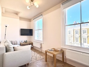 Ein Wohnzimmer mit einer Couch, einem Fernseher, einem Tisch mit Zeitschriften und einem großen Fenster mit Blick auf Gebäude.