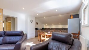 A modern living room with leather sofas, a dining area with wooden chairs, a kitchen with a built-in oven, and a large window. - Gite du Nant des l'Alouy (ESSERTS BLAY)