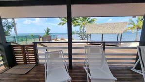 Una terrazza coperta con due poltrone bianche, pavimento in legno, ombrellone di paglia e vista sull'oceano e sulle palme.