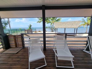 Eine überdachte Terrasse mit zwei weißen Liegestühlen, Holzboden, Sonnenschirm aus Schilf und Blick auf das Meer und Palmen.