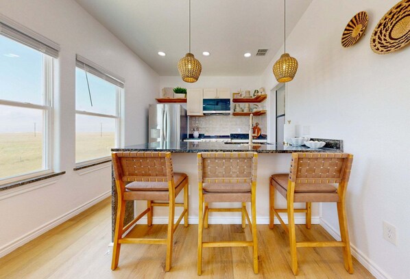 A kitchen with a central island, three wicker chairs, pendant lights, and a view through large windows. - WPB142: Walk to the Beach, Pet Friendly, Rooftop Terrace (Port Aransas)