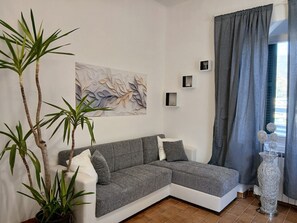 A modern living room with a grey sectional sofa, a large abstract painting, a potted plant, a white vase, and a grey curtain.