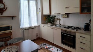 Kompakti keittiö, jossa on liesi, uuni ja pesuallas, ruokapöytä tuoleineen sekä ikkuna, jossa on verhot.