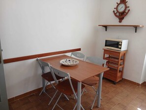 A small dining area with a wooden table, two chairs, a bowl, a microwave on a wooden stand, and a wall clock.