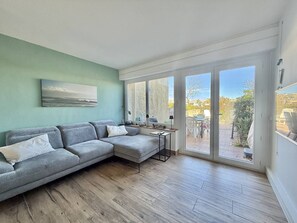 Ein Wohnzimmer mit einer grauen Eckcouch, einem kleinen Tisch und einem Fenster mit Blick auf eine Terrasse.