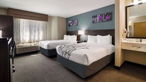 A hotel room with two beds, a desk, a chair, a window with curtains, a wall-mounted TV, and a bathroom with a mirror and toiletries.