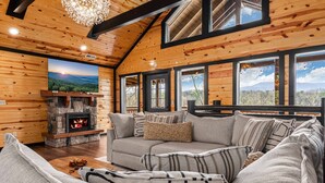 A cozy living room with a stone fireplace, a large window with a mountain view, and a comfortable sofa with patterned cushions. - 2 Bowling Lanes | 8BR Smoky Mtn Lodge | Hot Tub + 85° Pool + Arcade | Sleeps 22 (Sevierville)