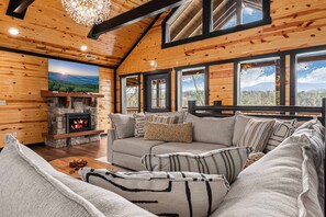 A cozy living room with a stone fireplace, a large window with a mountain view, and a comfortable sofa with patterned cushions.