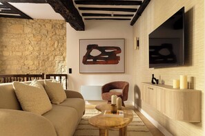 A cozy living room with a stone wall, a wooden sofa, a round coffee table, a brown armchair, and a mounted TV. - Premium Souplex a/c 1br/4p - Saint-germain (Paris)