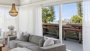 Un saló modern amb un sofà gris, coixins blancs, una làmpada penjant de vimet i vista a la platja a través de grans finestres.
