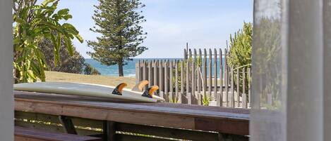 Una panca di legno con un surfboard appoggiato, con vista su una spiaggia, una recinzione di legno e alberi.
