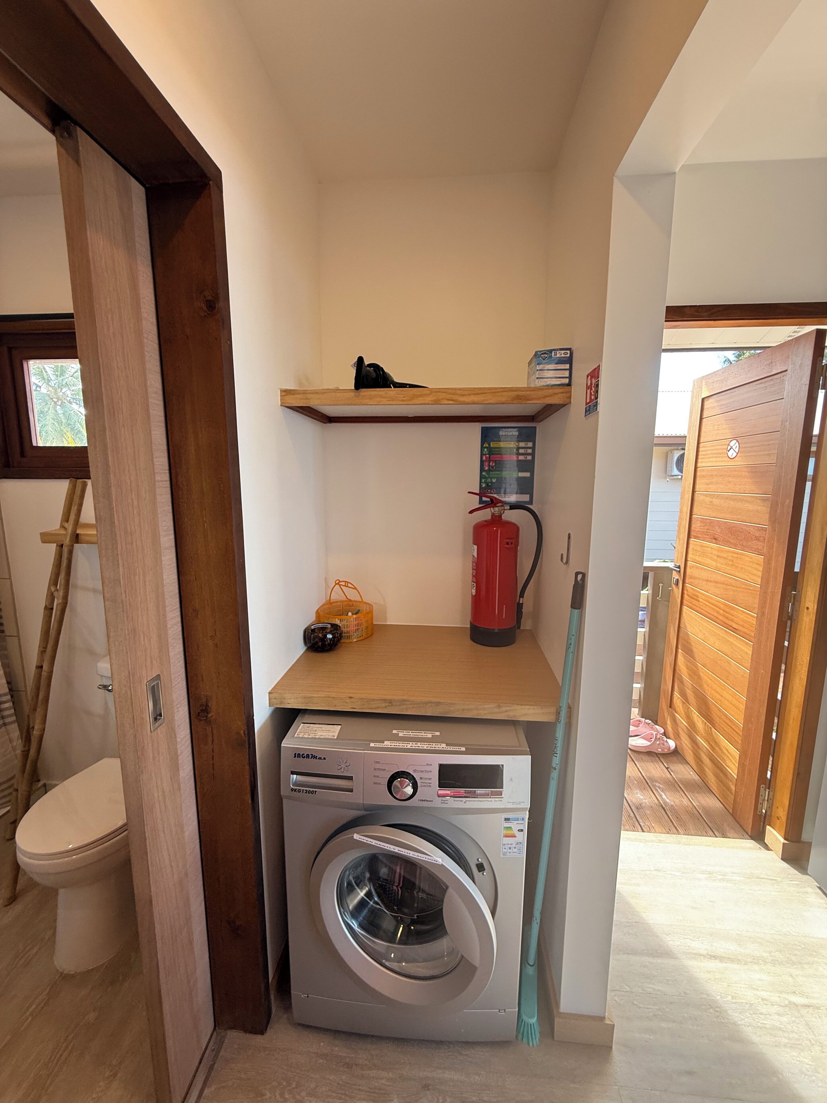 A compact laundry area with a washing machine, a fire extinguisher, and a shelf with a basket and a pair of shoes.