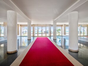 Eine großzügige Lobby mit rotem Teppich, hohen Säulen und Blick auf Strand und Meer durch Glastüren.