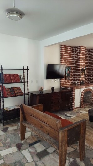 A living room with a brick fireplace, a flat-screen TV mounted above it, a wooden bench, and a bookshelf with books. - A paradise among mountains, 1 hour from Rio, for up to 60 people. (Guapimirim)