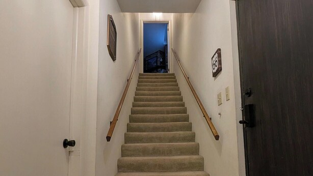 Una escalera alfombrada con barandilla, una puerta con placa de número y un artefacto de iluminación en el techo.