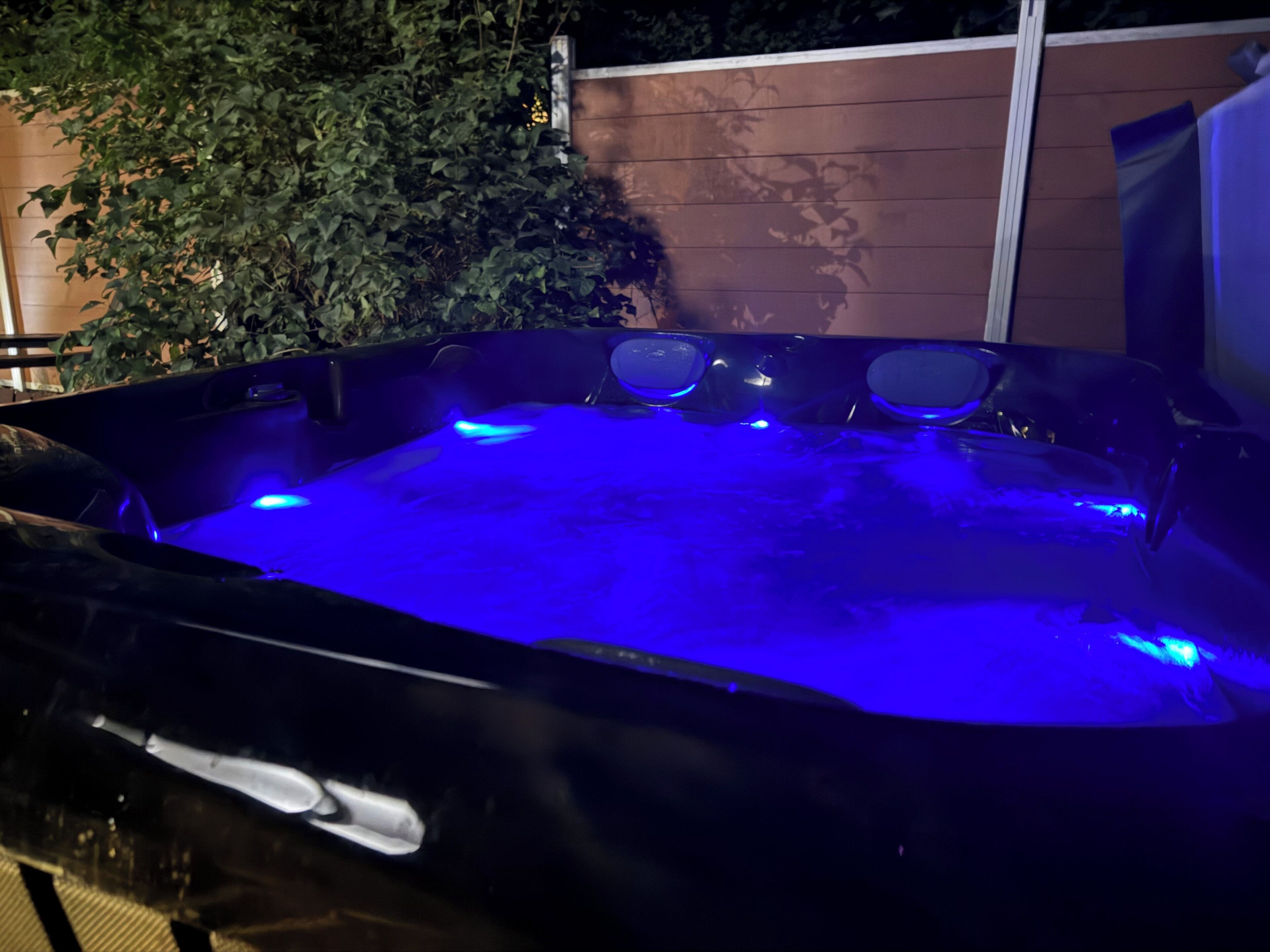 Un jacuzzi con iluminación azul, rodeado de follaje y una cerca de madera.