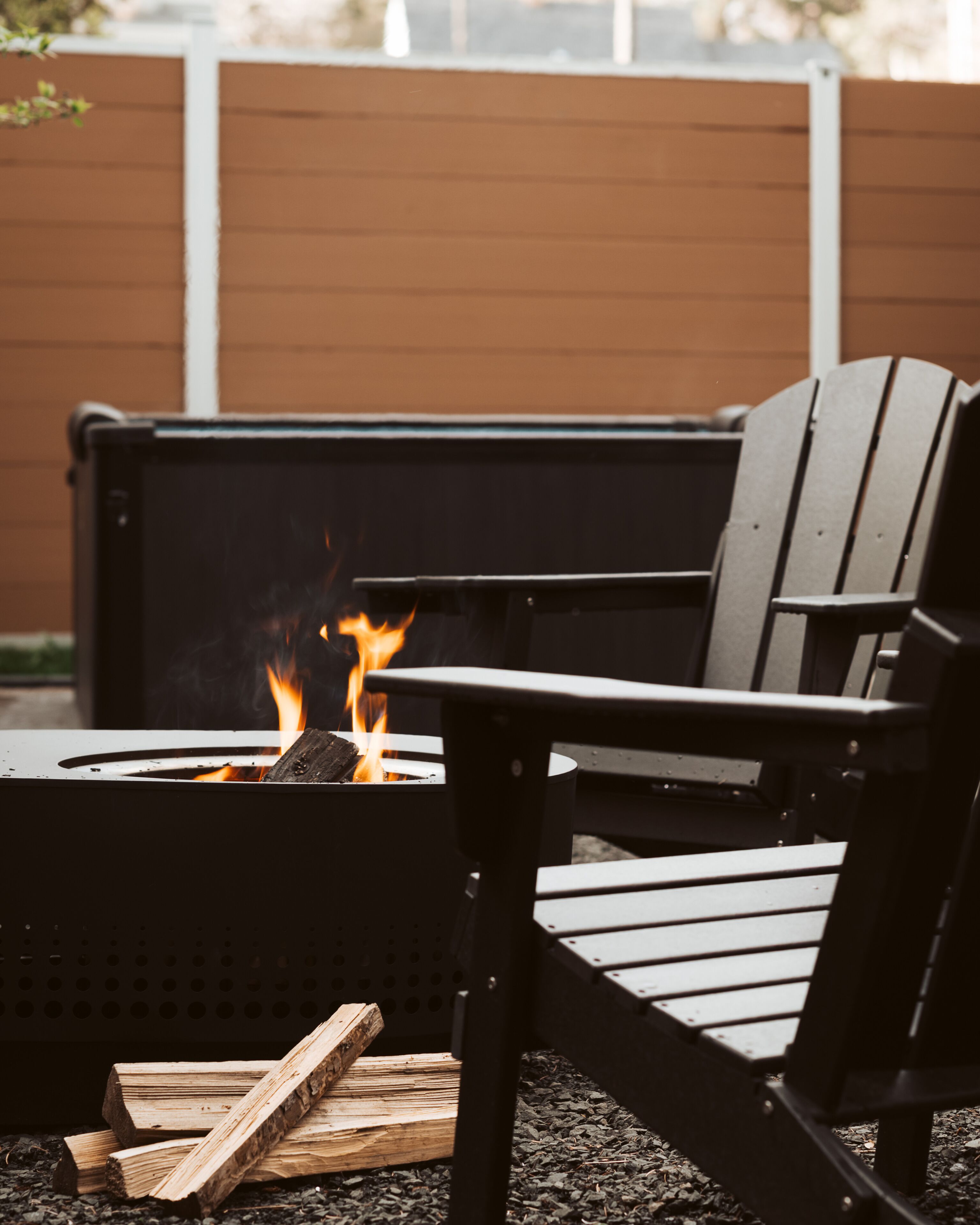 A black Adirondack chair in front of a fire pit with burning logs.
