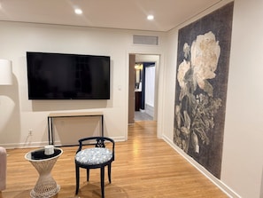 Un passadís amb un televisor de pantalla plana fixat a la paret, una taula petita amb una cadira, i un quadre gran amb motius florals.