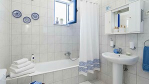 A bathroom with a bathtub, a sink, and a window with a curtain. There are decorative plates on the wall and a towel rack with towels. - Pool Villa Overlooking the Sea (Rhodes)