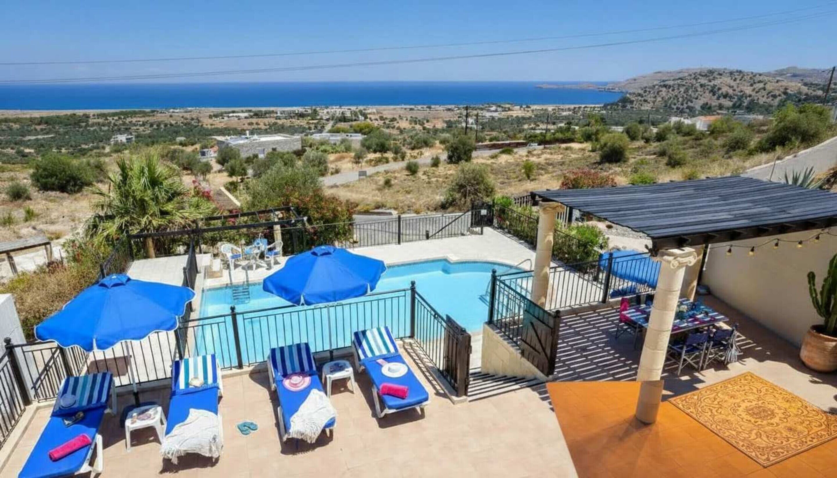A pool area with lounge chairs and umbrellas, a view of the sea, and a hilly landscape in the background.